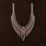 Straight-on view of long necklace on dark brown background, central floral pendant with red stones