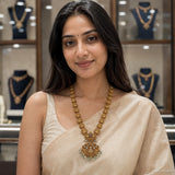 Woman in a beige saree wearing a long antique gold-tone necklace with a floral pendant and bead drops