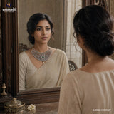 Woman looking into an ornate mirror wearing a beige saree and a green stone studded choker necklace with pearl drop